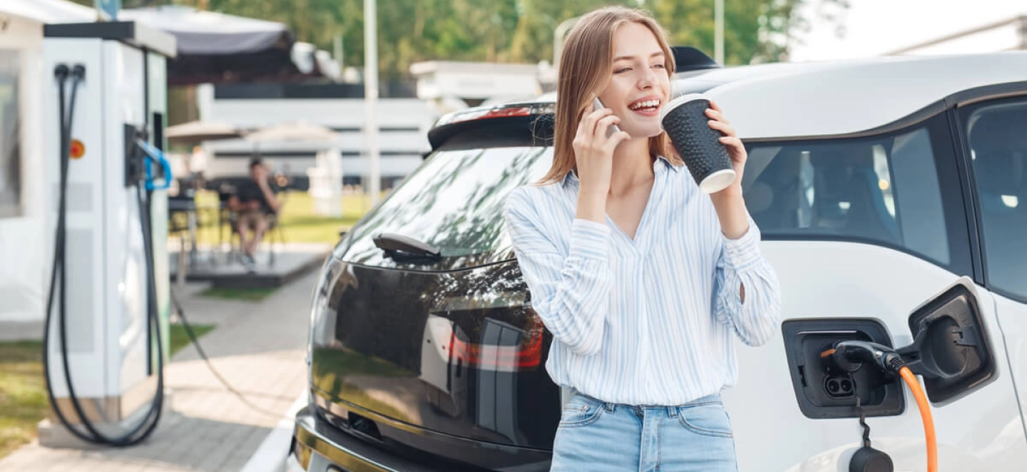 Eine junge Frau lädt ihr E-Auto an einer öffentlichen Ladestation und trinkt Kaffee.