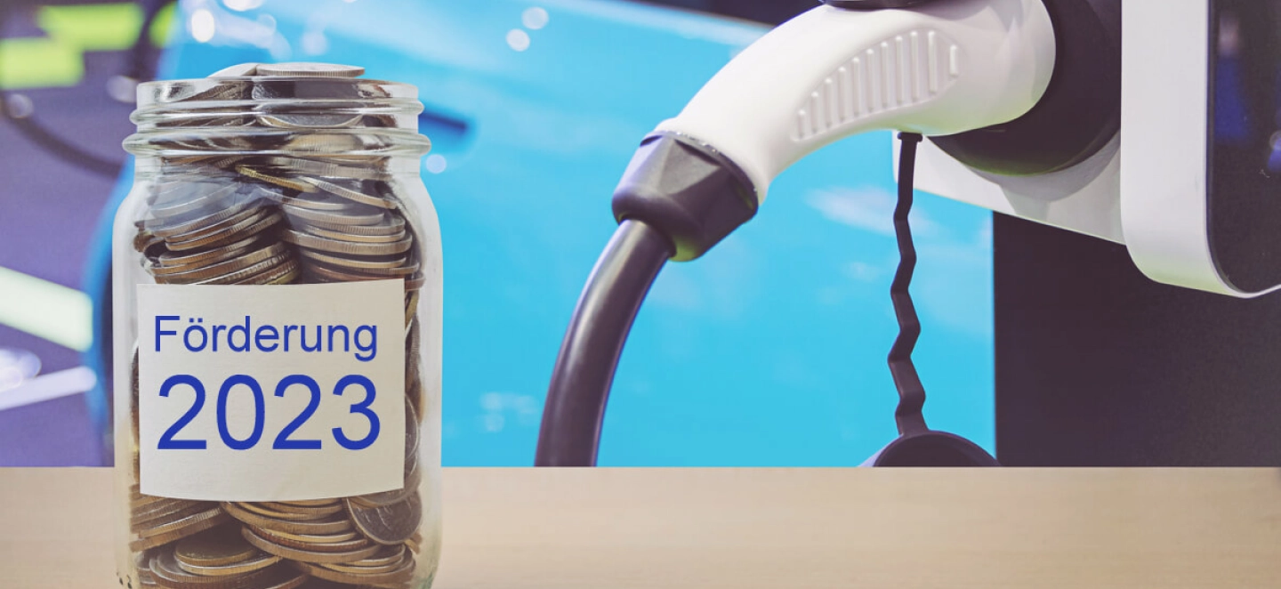 Vorn im Bild steht ein Glas mit Geld und der Aufschrift "Förderung 2023" und im Hintergrund wird ein Elektroauto mit Strom betankt.