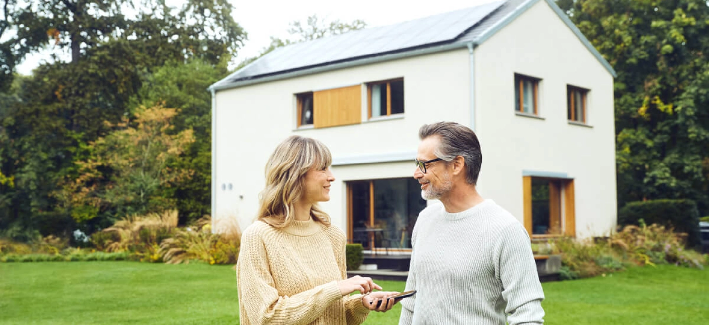 Ein Mann und eine Frau stehen im Garten und reden während im Hintergrund ihr Haus mit Solaranlage zu sehen ist.