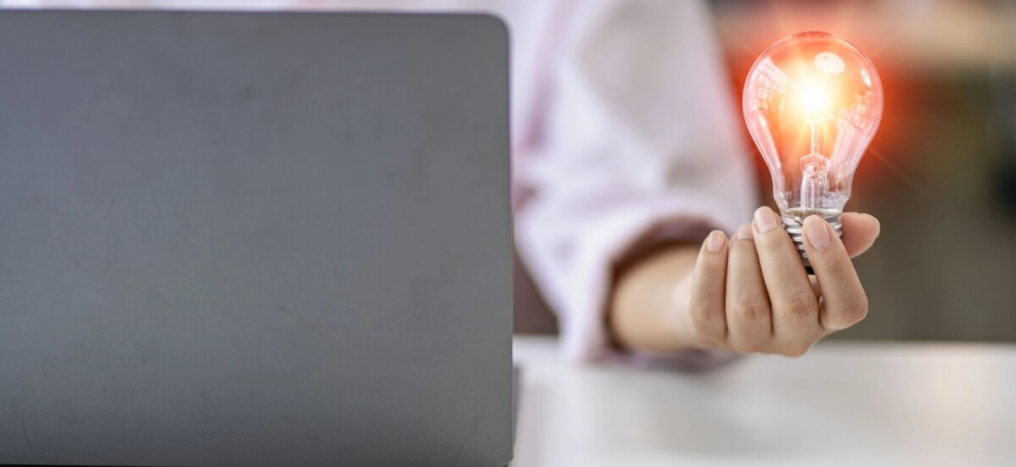 Eine Person sitzt vor einem Laptop und hält eine leuchtende Glühbirne in der Hand.