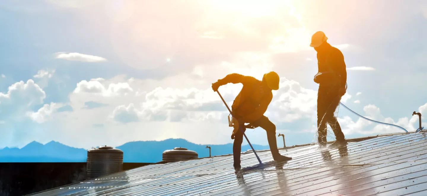 Zwei Arbeiter putzen Photovoltaik-Module auf einem Dach mit Sonne im Hintergrund.