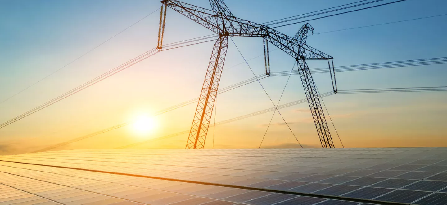 Im Vordergrund ist eine Solaranlage und im Hintergrund ist ein öffentlicher Hochspannungsmast und am Horizont ist der Sonnenaufgang zu sehen.