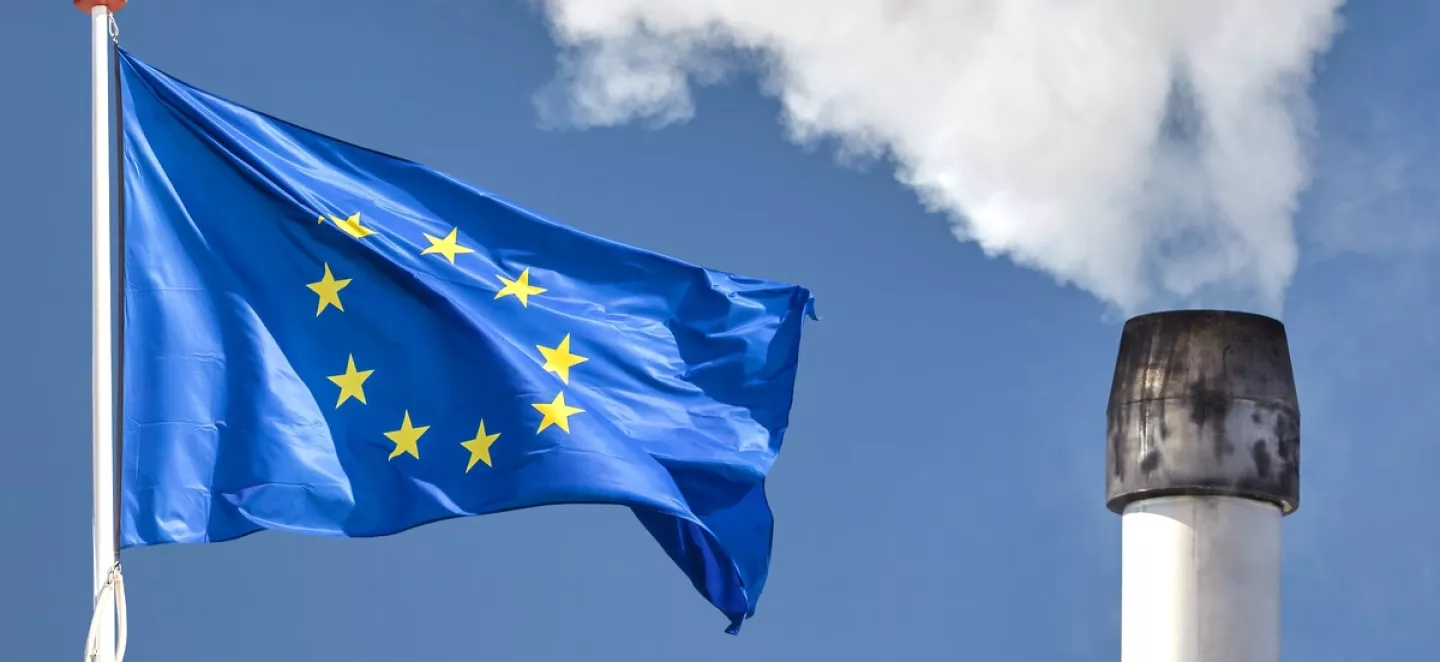 Links weht die europäische Flagge und rechts steht ein qualmender Industrie-Schornstein.