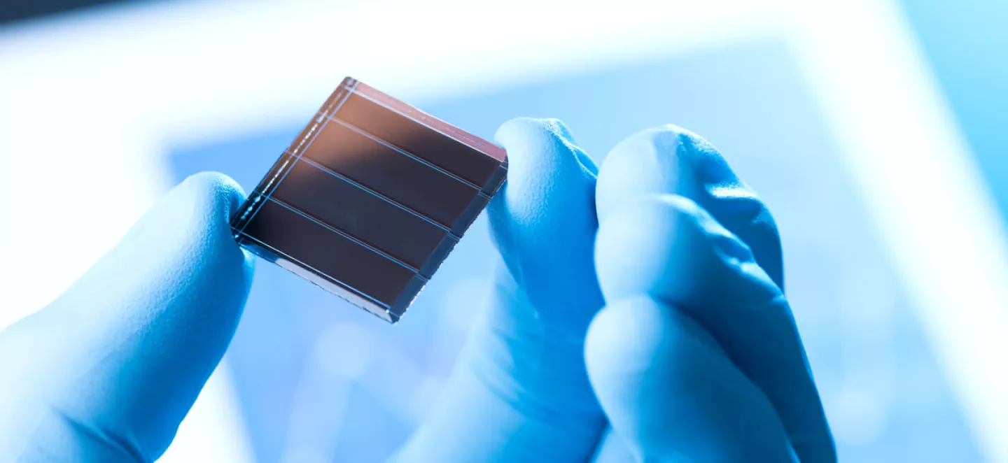 Eine Solarzelle zwischen Daumen und Zeigefinger einer Hand, die einen blauen Gummihandschuh trägt. Die Person ist in einem Labor für Solarzellen.