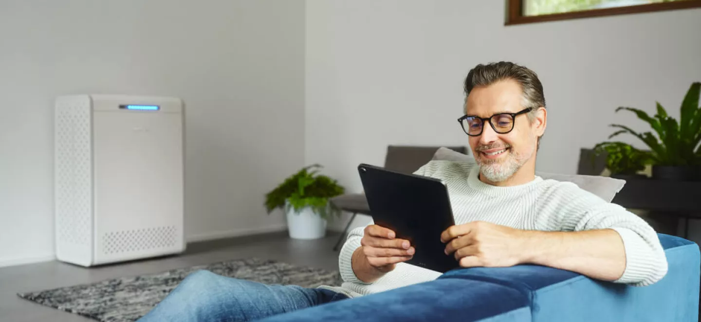 Ein Mann sitzt auf einem Sofa im Wohnzimmer, während im Hintergrund sein neu gekaufter Stromspeicher steht.