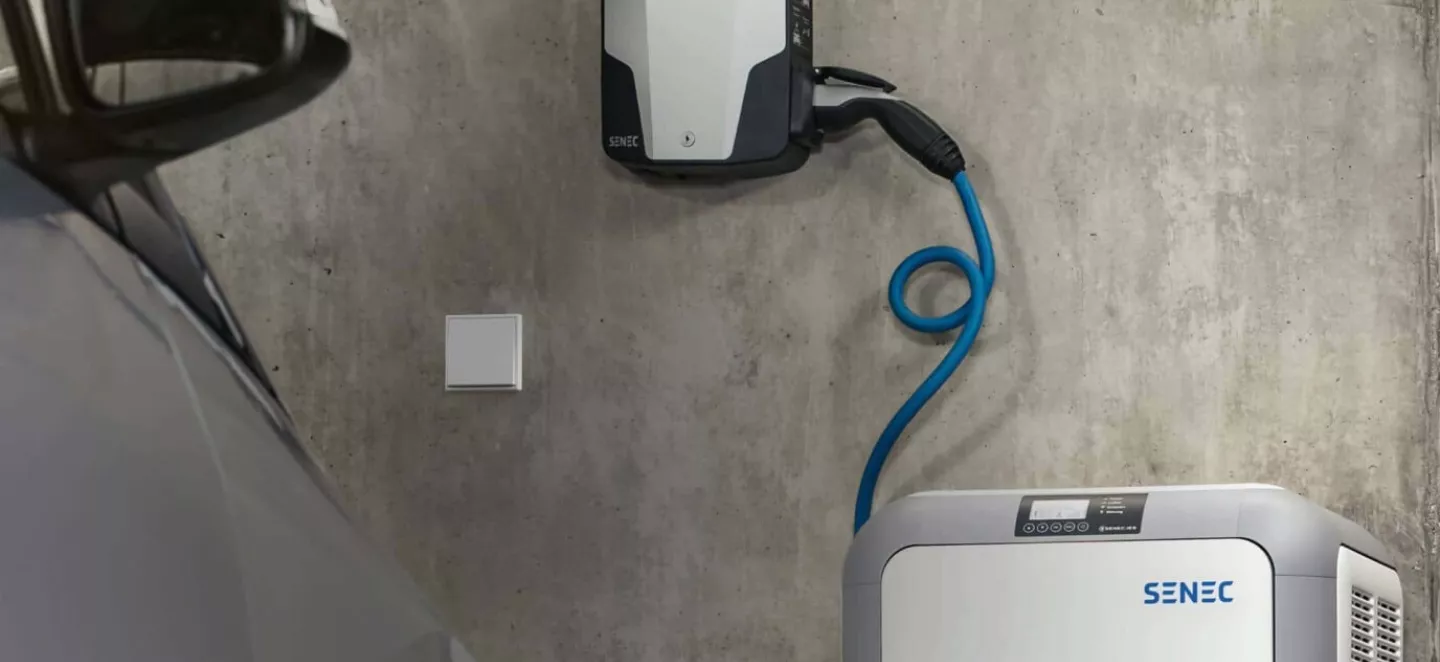 Eine moderne Wallbox hängt an der Wand. Darunter steht ein verbundener Stromspeicher. Vorn links ist die Ecke eines Autos zu sehen.