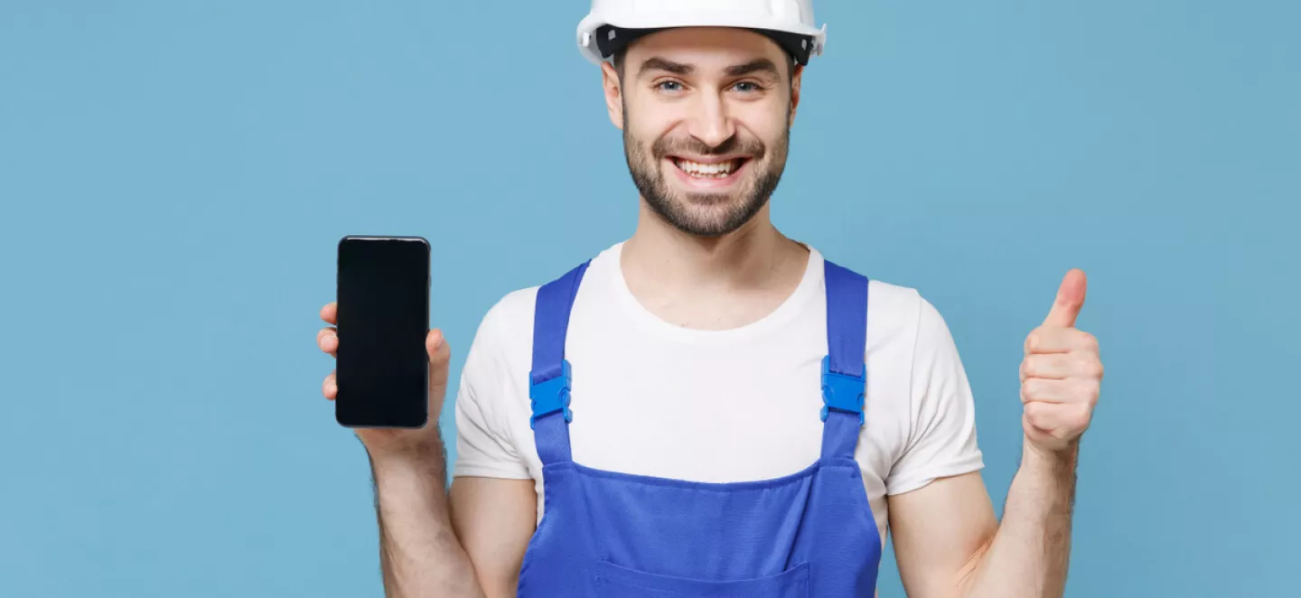 Handwerker mit weißem Helm und blauer Latzhose hält lächelnd sein Smartphone hoch und zeigt einen Daumen nach oben für Social-Media.