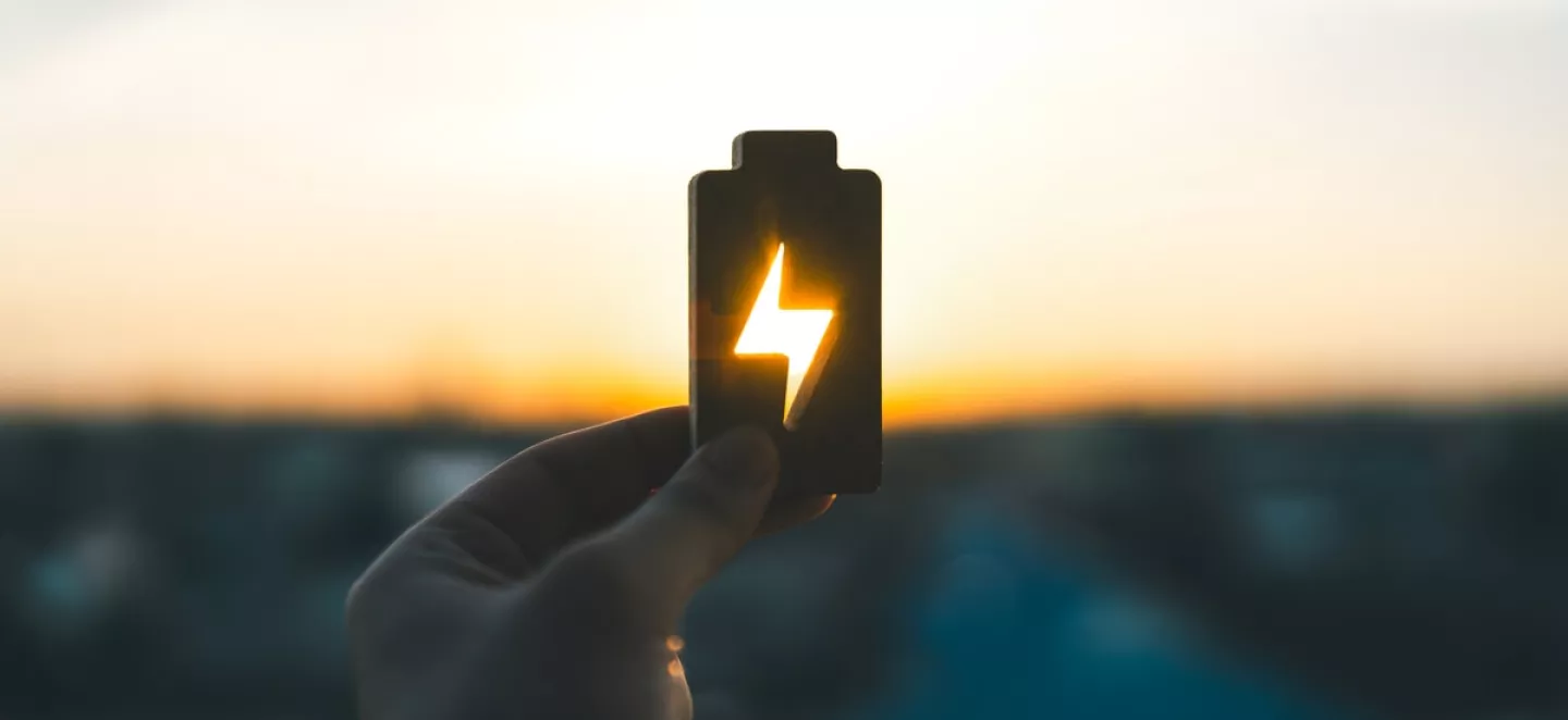 Eine Hand hält dem Umriss einer Batterie in den Himmel, während die untergehende Sonne durch das Strom-Symbol scheint und leuchtet.