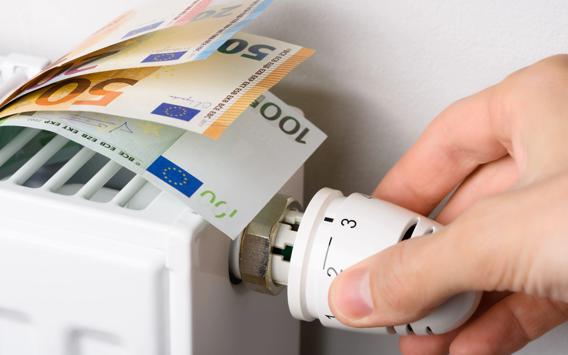 Auf einer Heizung liegen Euro-Scheine, während eine Hand das Thermostat dreht.