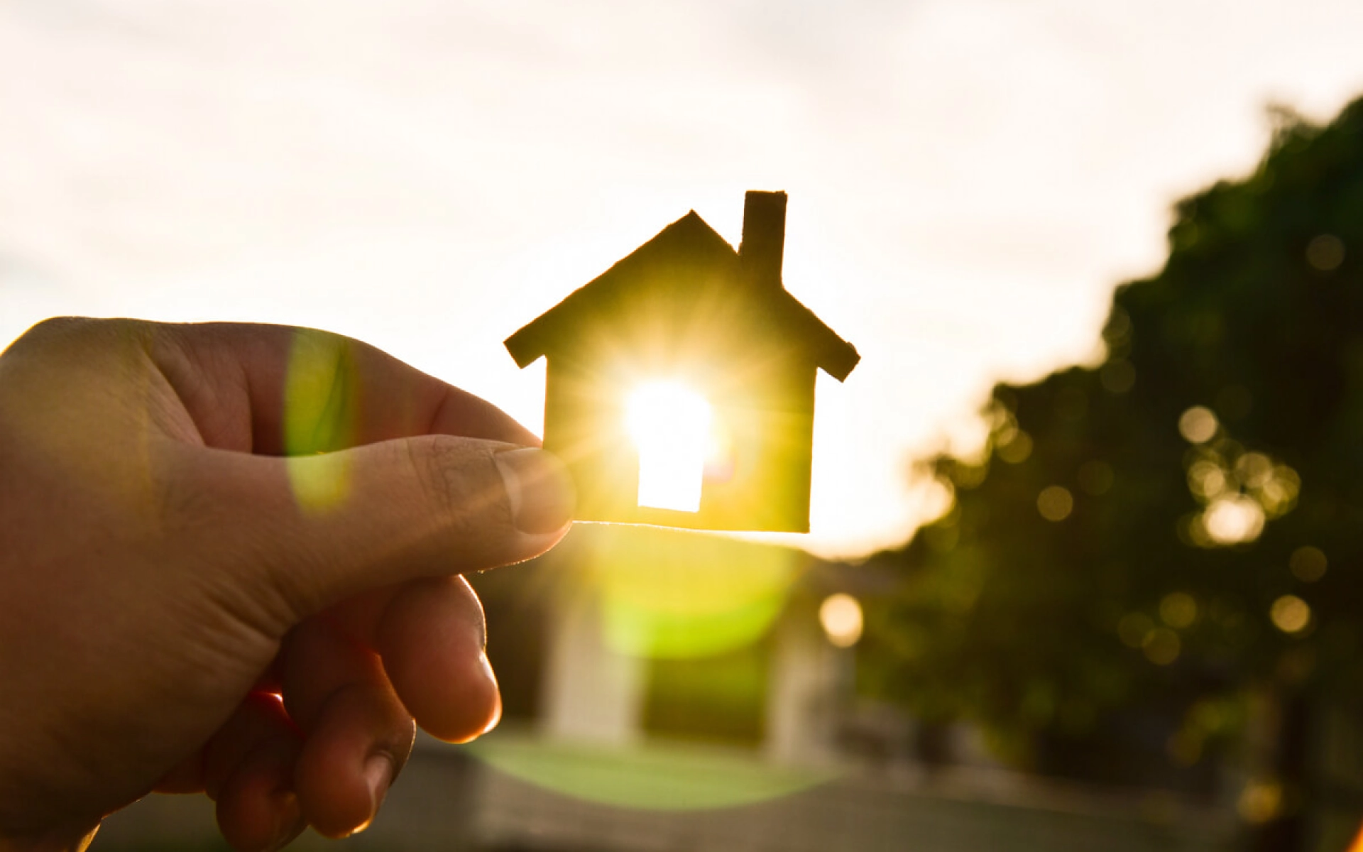 Eine Hand hält ein kleines Haus aus Holz gegen die Sonne und die Sonne scheint durch ein Fenster des kleinen Hauses. 