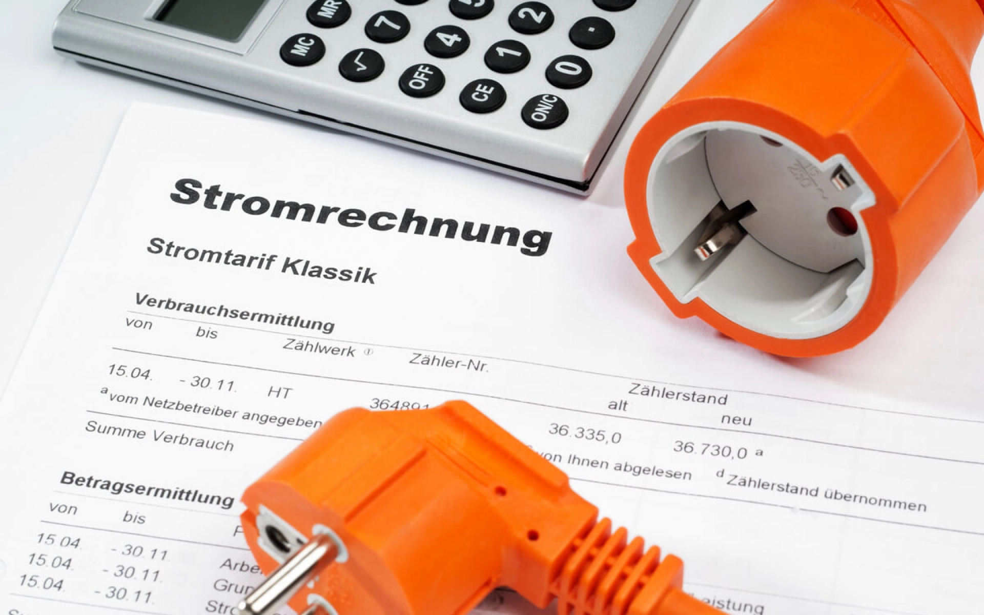 Auf einem Tisch liegt ein Taschenrechner, mehrere Seiten einer Stromrechnung und eine Steckdosen mit Stecker und Stromkabel.