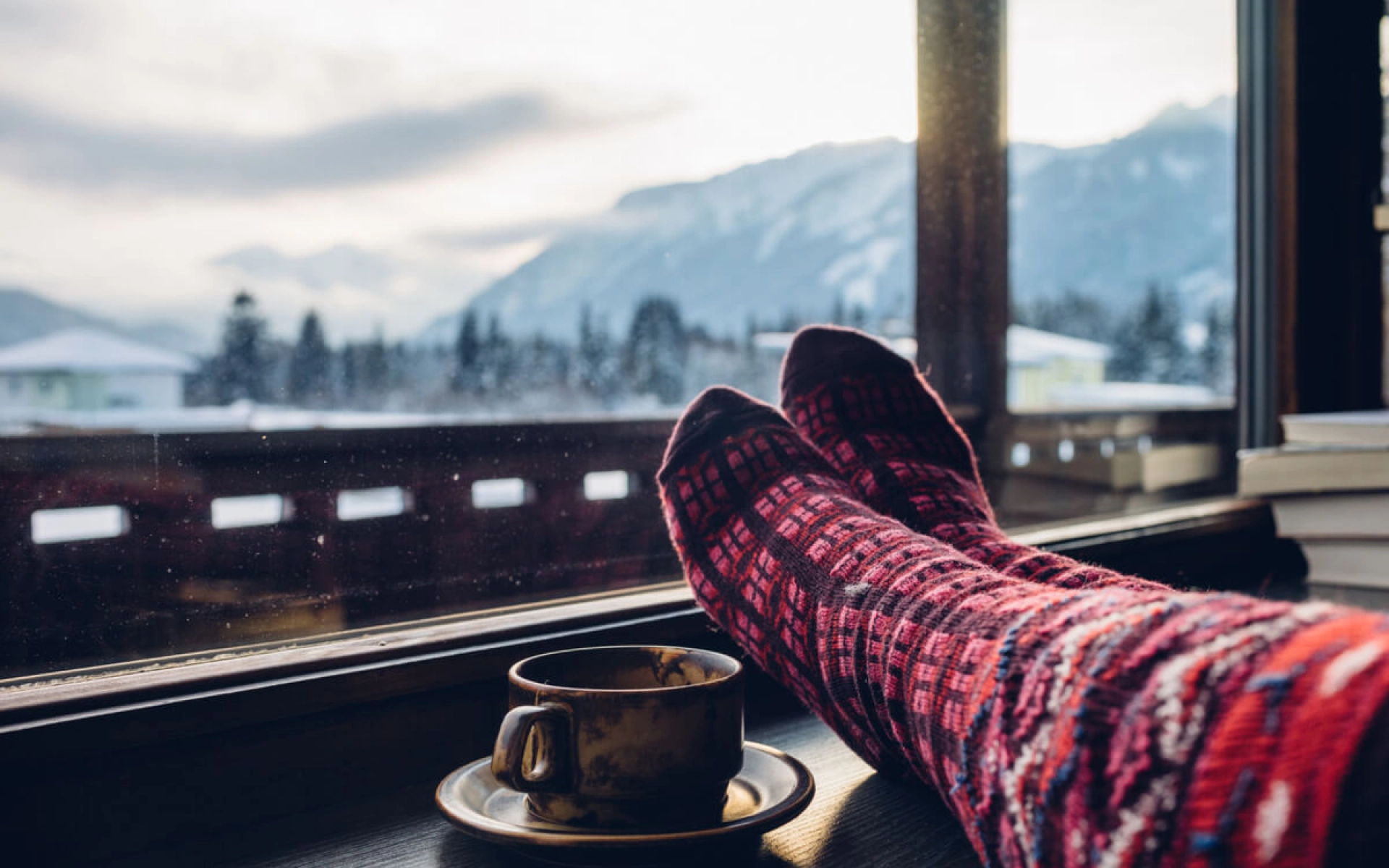 Warme Wintersocken und eine Tasse Tee stehen auf einer Fensterbank und der Blick fällt hinaus auf eine Schneelandschaft.