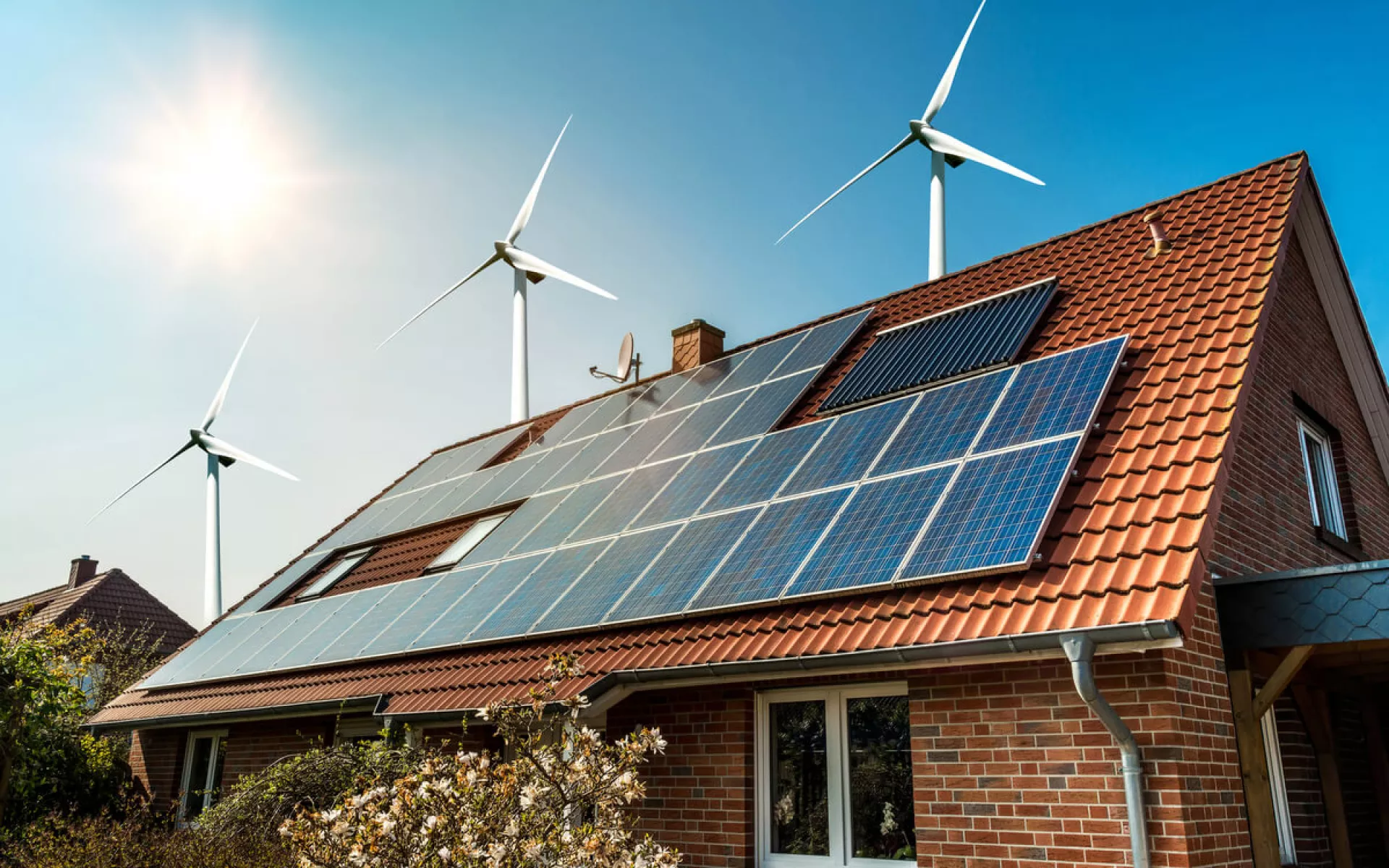 Ein Hausdach, das Sonnenkollektoren installiert hat. Im Hintergrund sind Windkrafträder und blauer Himmel.