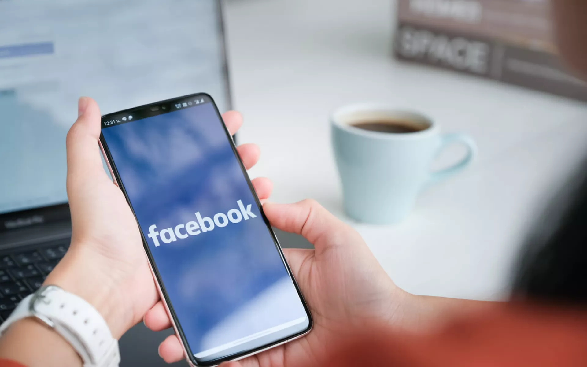 Zwei Hände halten ein Smartphone über dem Schreibtisch, auf dem Facebook geöffnet ist. Im Hintergrund steht ein aufgeklappter Laptop und eine Tasse Kaffee.