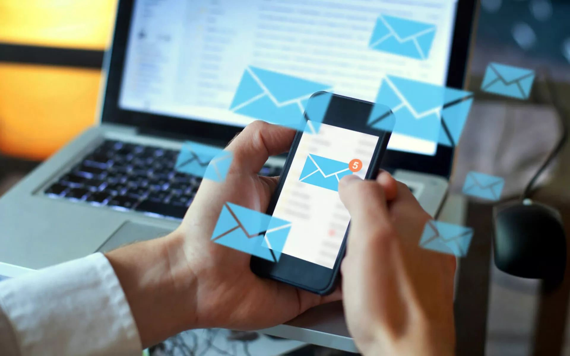 Zwei Hände halten ein Smartphone, auf dem mehrere Newsletter-Nachrichten ankommen. Im Hintergrund ist ein offener Laptop mit E-Mails.