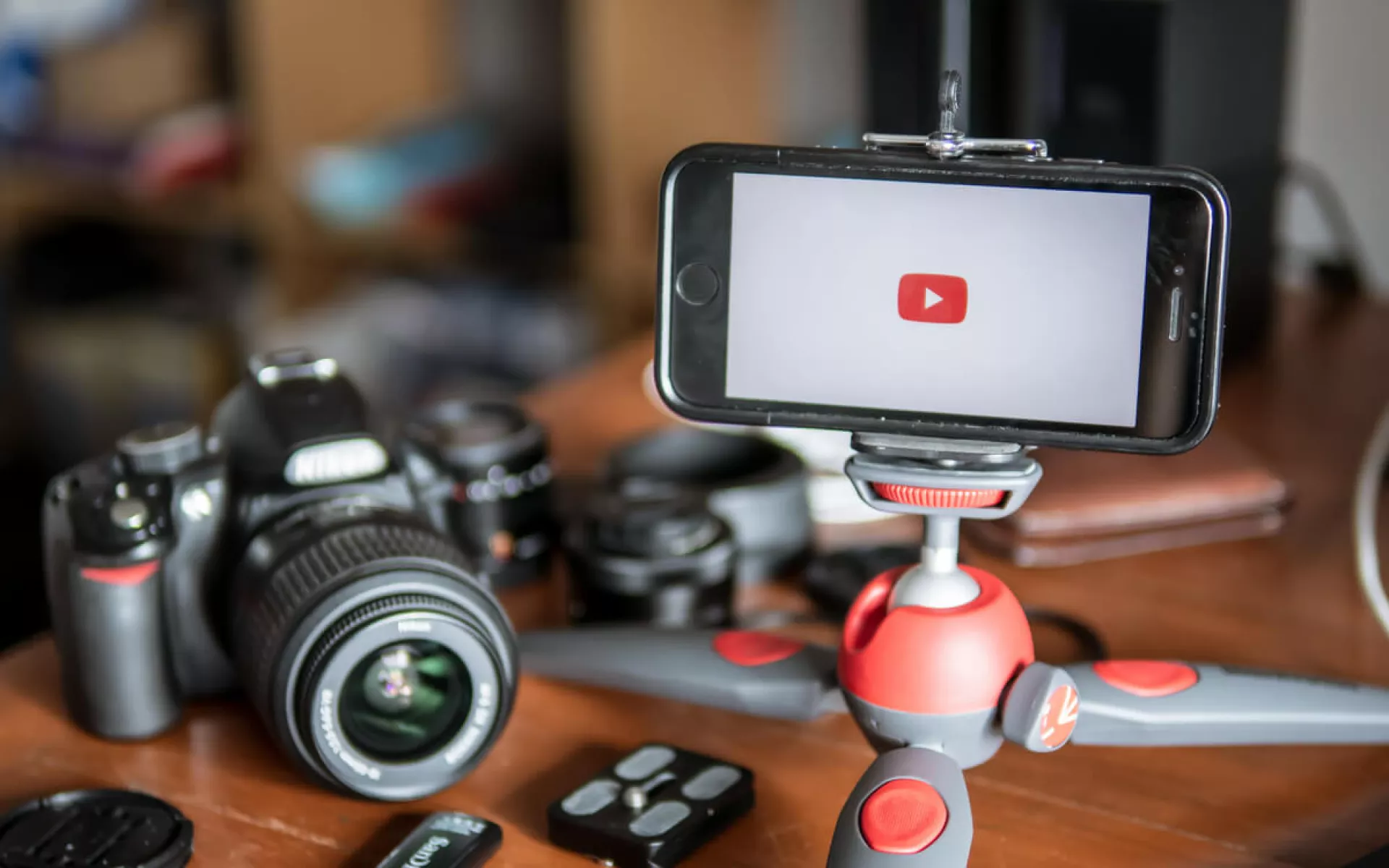 Auf einem Tisch liegt Equipment zum Erstellen von Youtube-Videos. Eine Kamera mit Objektiv, ein Smartphone mit drehbarem Ständer und ein Speicherstick mit weiteren Objektiven.