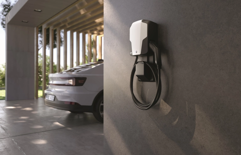 Eine moderne Wallbox hängt an einer Hauswand eines Privathauses und im Hintergrund steht ein weißes Elektroauto zum Laden bereit.
