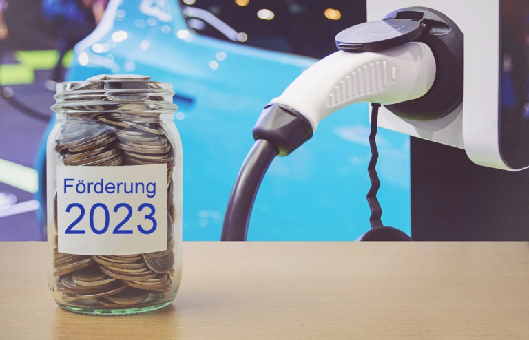 Vorn im Bild steht ein Glas mit Geld und der Aufschrift "Förderung 2023" und im Hintergrund wird ein Elektroauto mit Strom betankt.