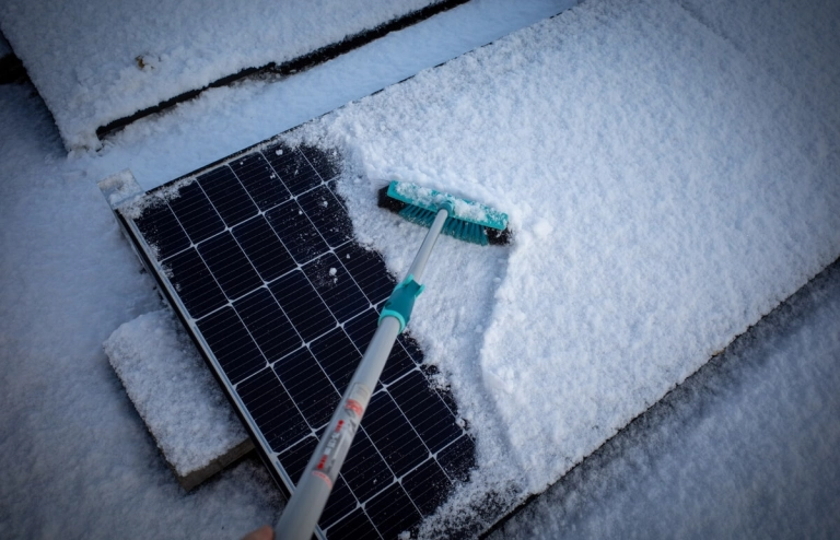 Schneebedeckte Solarmodule werden von einem Besen von Schnee befreit, sodass sie Strom erzeugen können. 