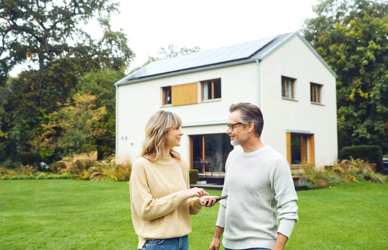 Ein Mann und eine Frau stehen im Garten und reden während im Hintergrund ihr Haus mit Solaranlage zu sehen ist. 