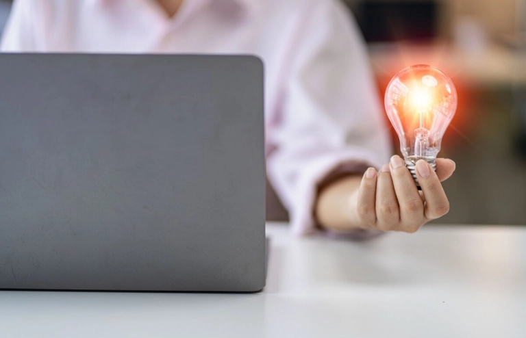 Eine Person sitzt vor einem Laptop und hält eine leuchtende Glühbirne in der Hand. 