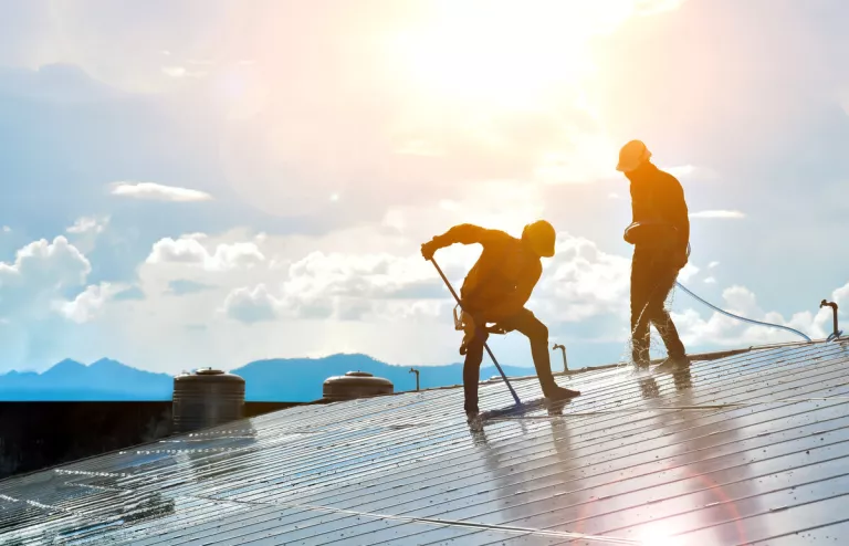 Zwei Arbeiter putzen Photovoltaik-Module auf einem Dach mit Sonne im Hintergrund. 