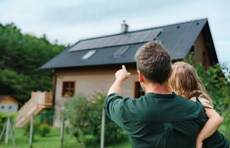 Ein Vater hat seine kleine Tochter auf dem Arm und zeigt mit dem Finger auf die Photovoltaik-Anlage auf dem Dach. 