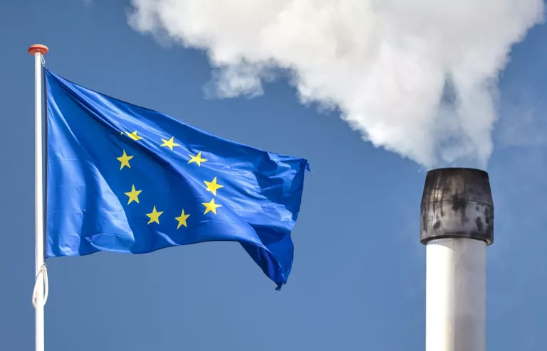 Links weht die europäische Flagge und rechts steht ein qualmender Industrie-Schornstein.