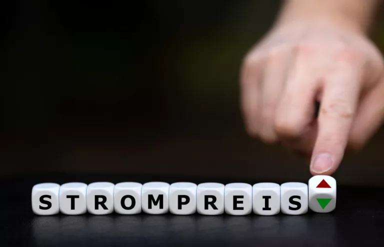 Im Vordergrund stehen Würfel mit Buchstaben, die das Wort Strompreis ergeben und eine Hand bewegt den letzten Würfel, auf dem ein grüner Pfeil nach oben und ein roter Pfeil nach unten zu sehen ist.