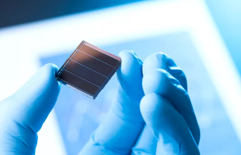 Eine Solarzelle zwischen Daumen und Zeigefinger einer Hand, die einen blauen Gummihandschuh trägt. Die Person ist in einem Labor für Solarzellen. 