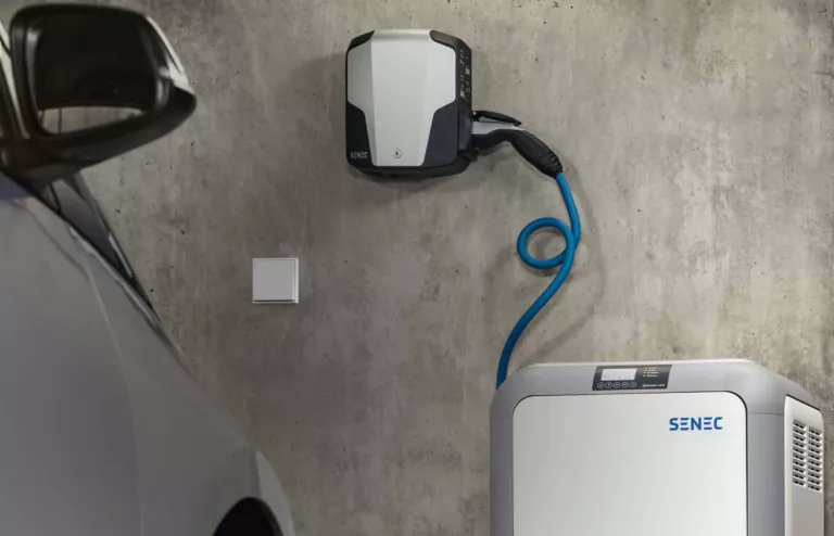 Eine moderne Wallbox hängt an der Wand. Darunter steht ein verbundener Stromspeicher. Vorn links ist die Ecke eines Autos zu sehen.