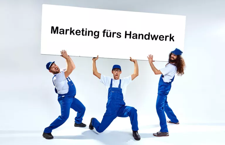 Drei Handwerker in blauen Latzhosen halten ein großes Schild hoch, auf dem Marketing für Handwerker steht.
