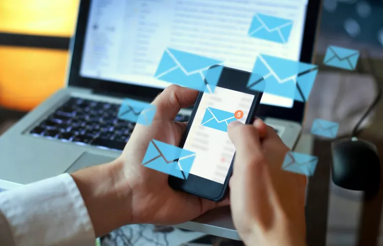 Zwei Hände halten ein Smartphone, auf dem mehrere Newsletter-Nachrichten ankommen. Im Hintergrund ist ein offener Laptop mit E-Mails.