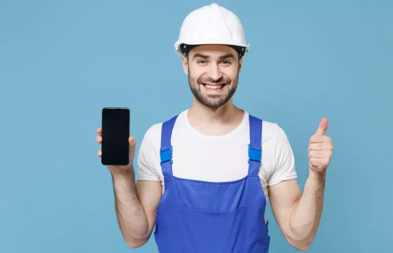 Handwerker mit weißem Helm und blauer Latzhose hält lächelnd sein Smartphone hoch und zeigt einen Daumen nach oben für Social-Media.