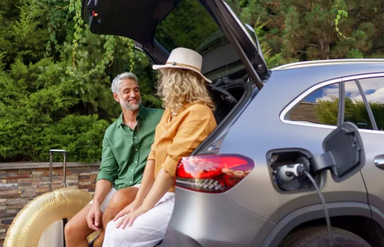 Ein Mann und eine Frau sitzen lachend im Kofferraum ihres E-Autos während das Ladekabel der Wallbox angeschlossen ist und das Fahrzeug auflädt.