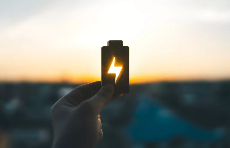 Eine Hand hält dem Umriss einer Batterie in den Himmel, während die untergehende Sonne durch das Strom-Symbol scheint und leuchtet.
