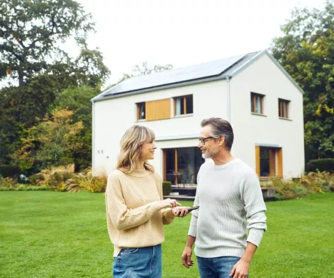 Ein Mann und eine Frau stehen im Garten und reden während im Hintergrund ihr Haus mit Solaranlage zu sehen ist.