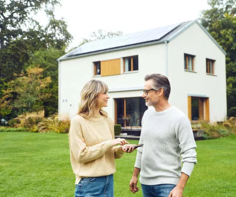 Eine Ehepaar im Gespräch und lachend. Im Hintergrund steht ihr Haus mit einer Solaranlage.