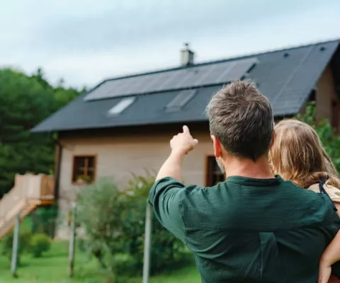 Ein Vater hat seine kleine Tochter auf dem Arm und zeigt mit dem Finger auf die Photovoltaik-Anlage auf dem Dach. 