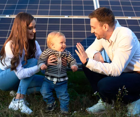Ein junger Vater und eine junge Mutter lachen und sitzen vor einem Solarpanel in der Sonne.