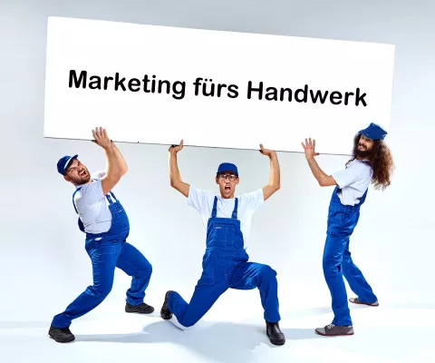 Drei Handwerker in blauen Latzhosen halten ein großes Schild hoch, auf dem Marketing für Handwerker steht.