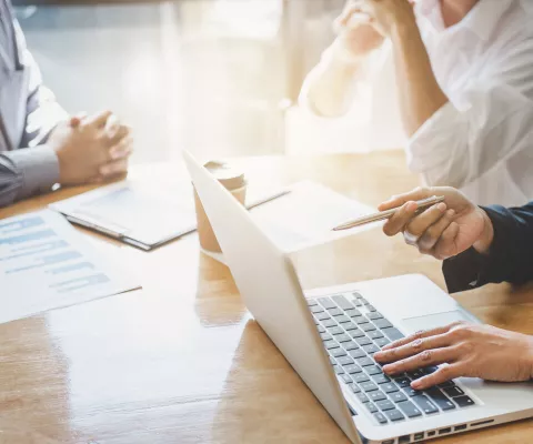 Drei Personen sitzen bei einem Beratungsgespräch. Es sind nur die Hände auf dem Tisch zu sehen. Dazwischen liegen Ausdrucke, Formulare und ein offener Laptop.