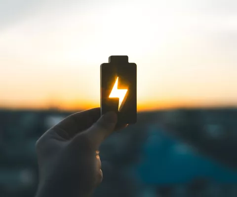 Eine Hand hält dem Umriss einer Batterie in den Himmel, während die untergehende Sonne durch das Strom-Symbol scheint und leuchtet.